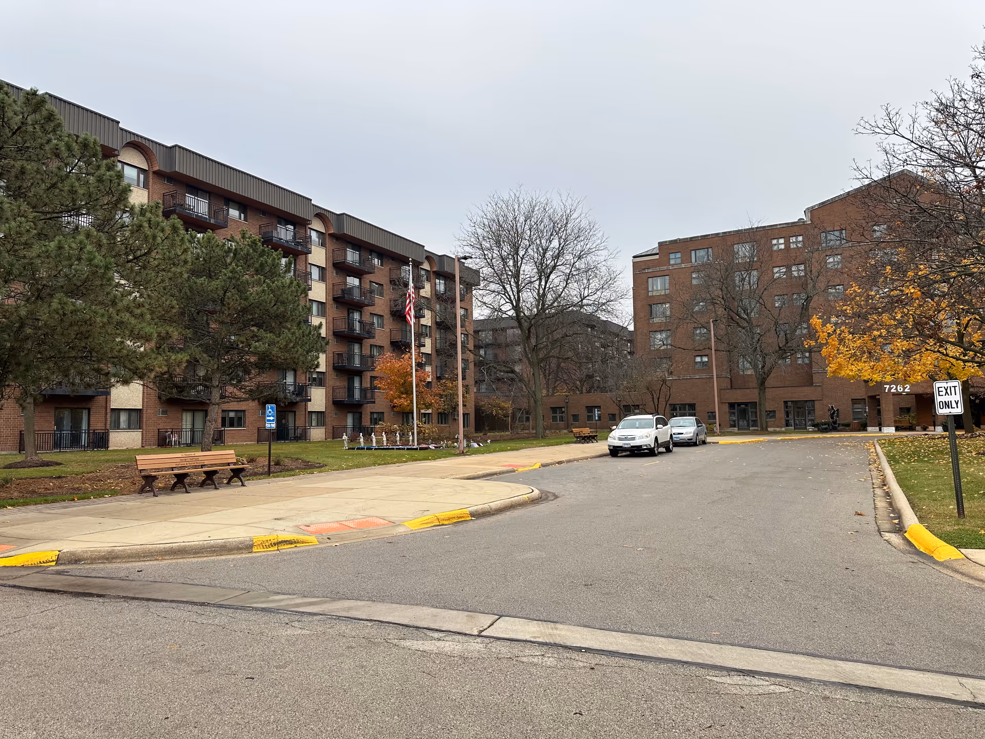 Exterior view of Ascension Living Resurrection Village showing a multi-story brick building with balconies, a driveway with two parked cars, benches, trees with autumn foliage, and an American flag on a flagpole.
