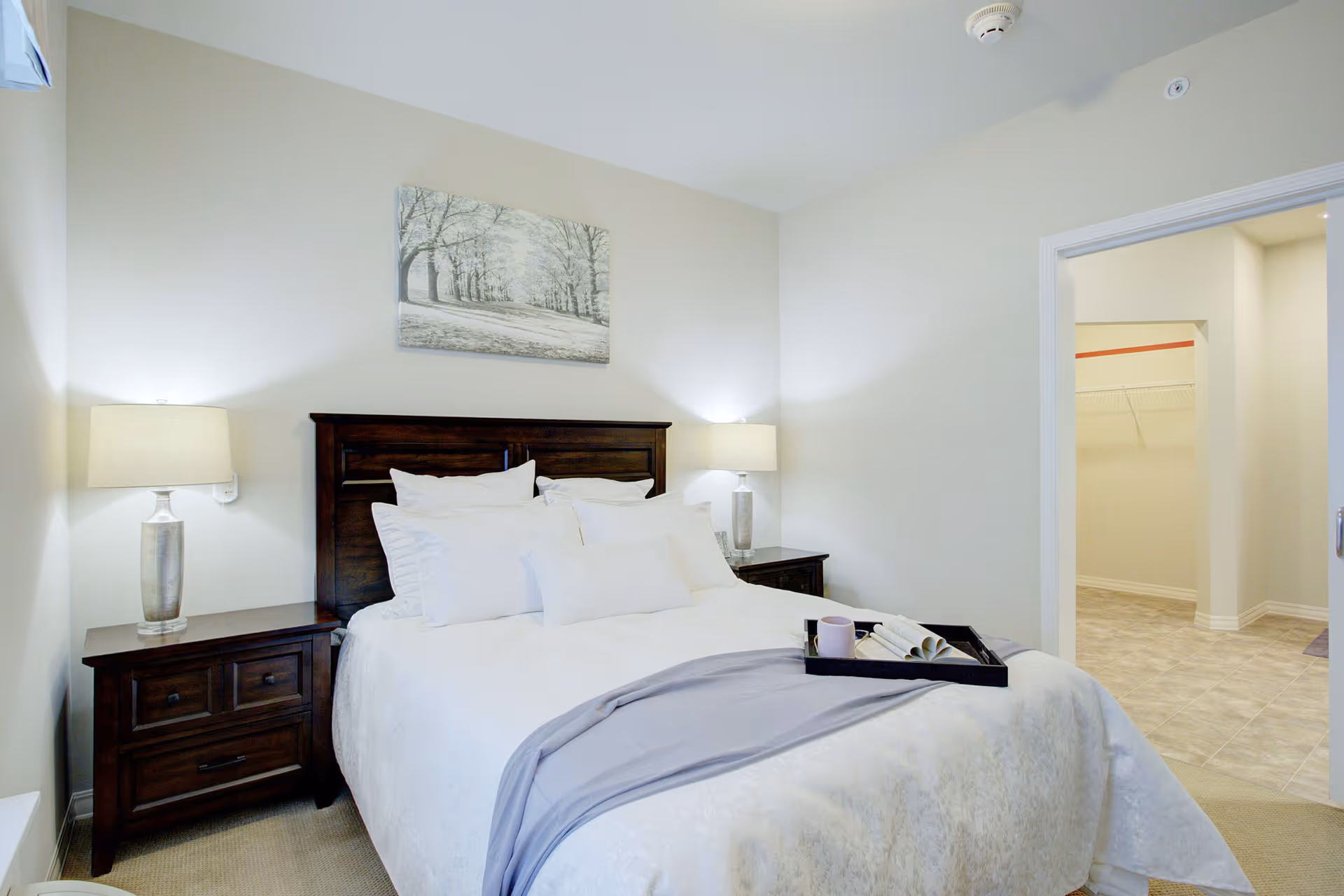 Neatly made bedroom with a wooden headboard, bedside tables and lamps, wall art, and an open door to a closet.