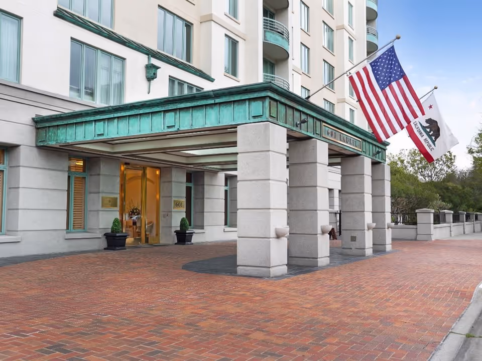 Covered entrance and porte-cochere of a multi-story building with an American and California flag and a brick driveway.