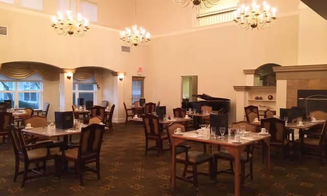 A spacious dining room with multiple tables set for a meal, featuring chairs with cushioned seats and backs. The room has high ceilings with chandeliers, large windows with draped curtains, a fireplace, and a grand piano in the corner.