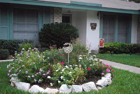 Front yard of a single-story house with a flower bed surrounded by white stones, green bushes, and a small lawn. The house has light blue shutters and a white front door with a small decorative sign next to it.