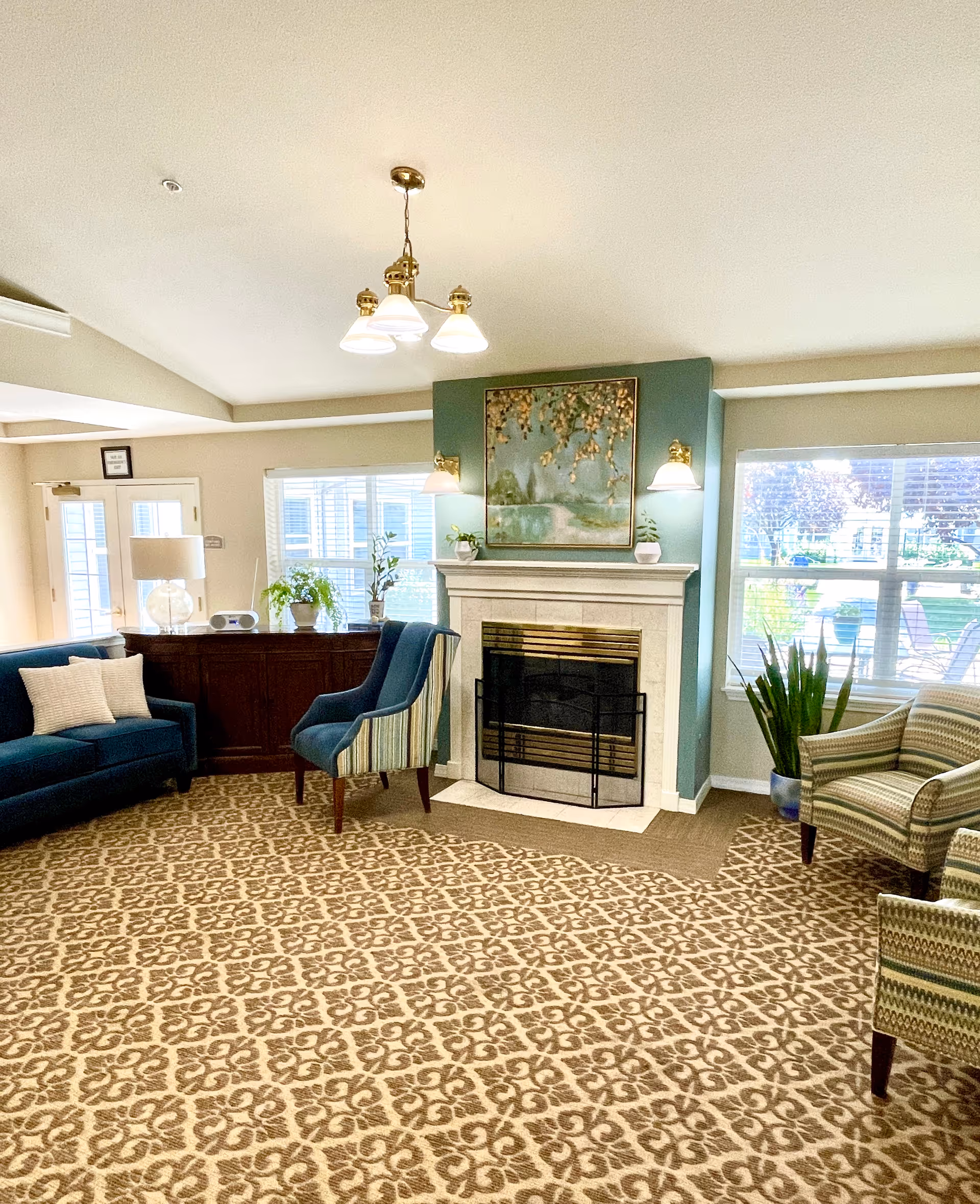 A cozy senior living facility common area with a patterned carpet, a white fireplace with a decorative screen, and a painting above it. The room has a blue-green accent wall behind the fireplace, two wall-mounted lamps, and several chairs including a dark blue sofa, a striped armchair, and two other patterned armchairs. There are large windows letting in natural light and a wooden cabinet with plants and a lamp on top.