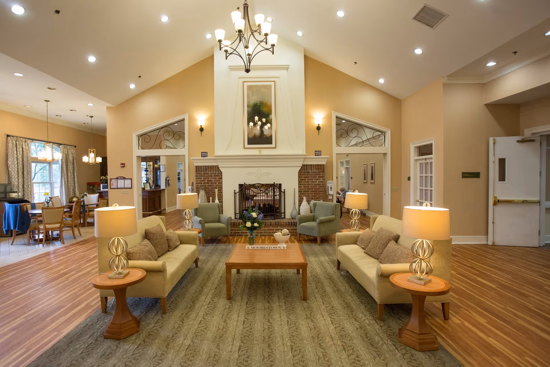 A spacious and well-lit living room area in a senior living facility with two beige sofas facing each other, two green armchairs, a wooden coffee table with a flower arrangement, and a large fireplace with a painting above it. The room has warm beige walls, wooden flooring, and several lamps providing soft lighting. In the background, there is a dining area with tables and chairs near large windows with curtains.