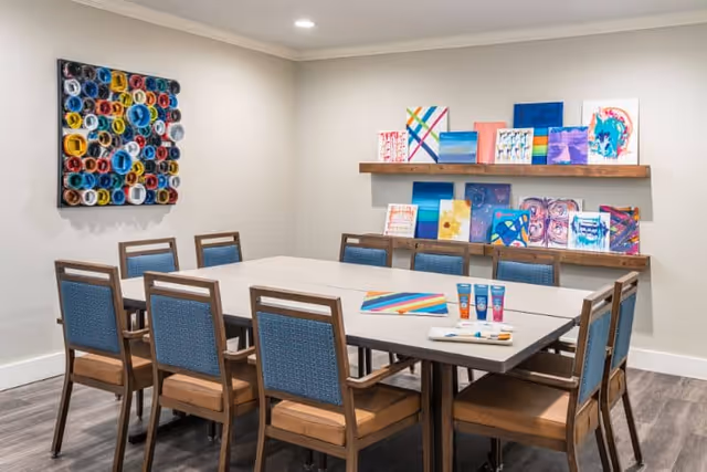A well-lit room with a large rectangular table surrounded by ten wooden chairs with blue upholstered backs and tan seats. On the table are several tubes of paint and a colorful painting. The walls are decorated with multiple colorful abstract paintings, some displayed on two wooden shelves and one mounted directly on the wall.