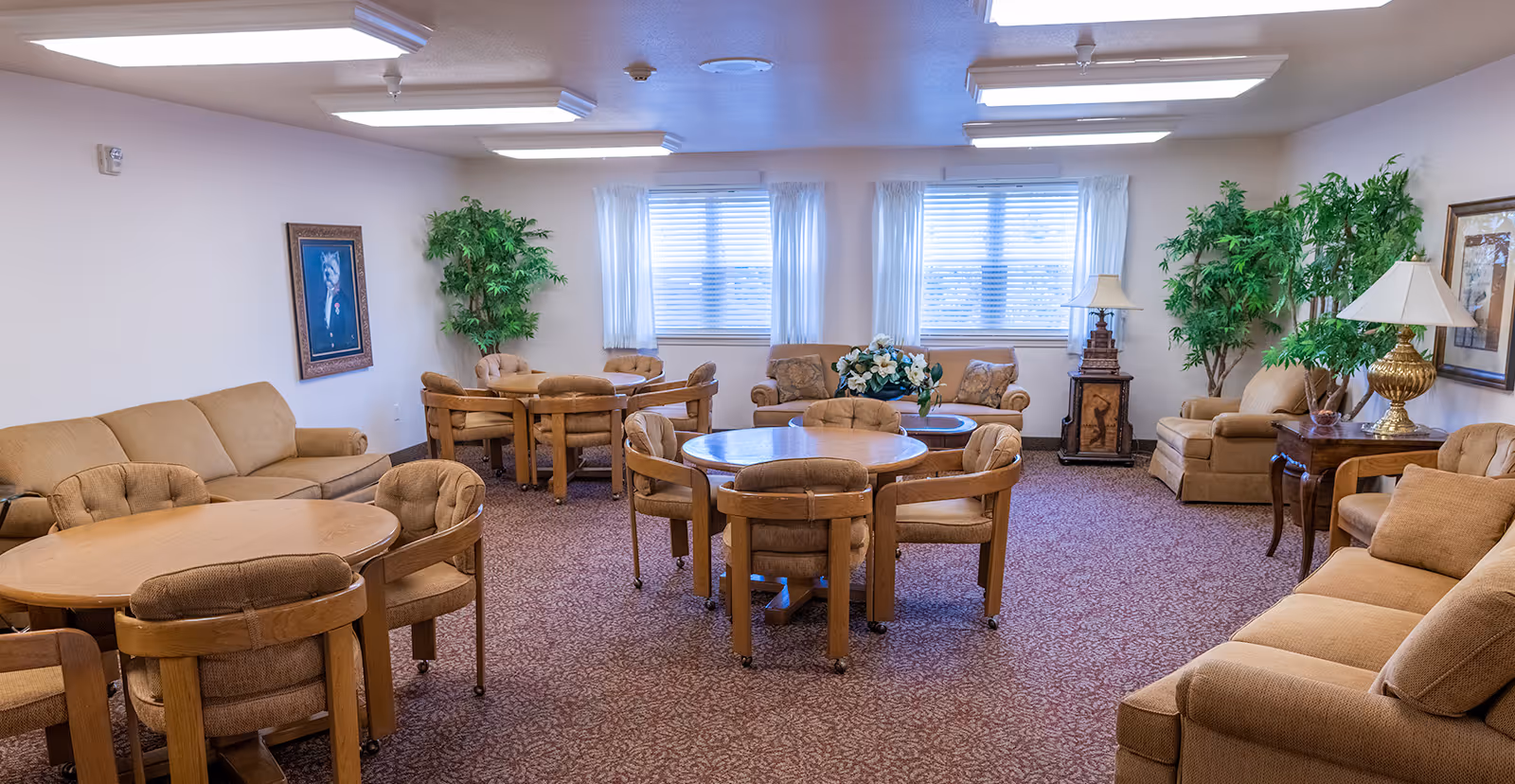 A spacious living room area with multiple round wooden tables surrounded by cushioned chairs. The room has beige sofas along the walls, two large windows with white curtains, green potted plants, framed artwork on the walls, and table lamps on side tables. The carpet is patterned in a reddish-brown color.