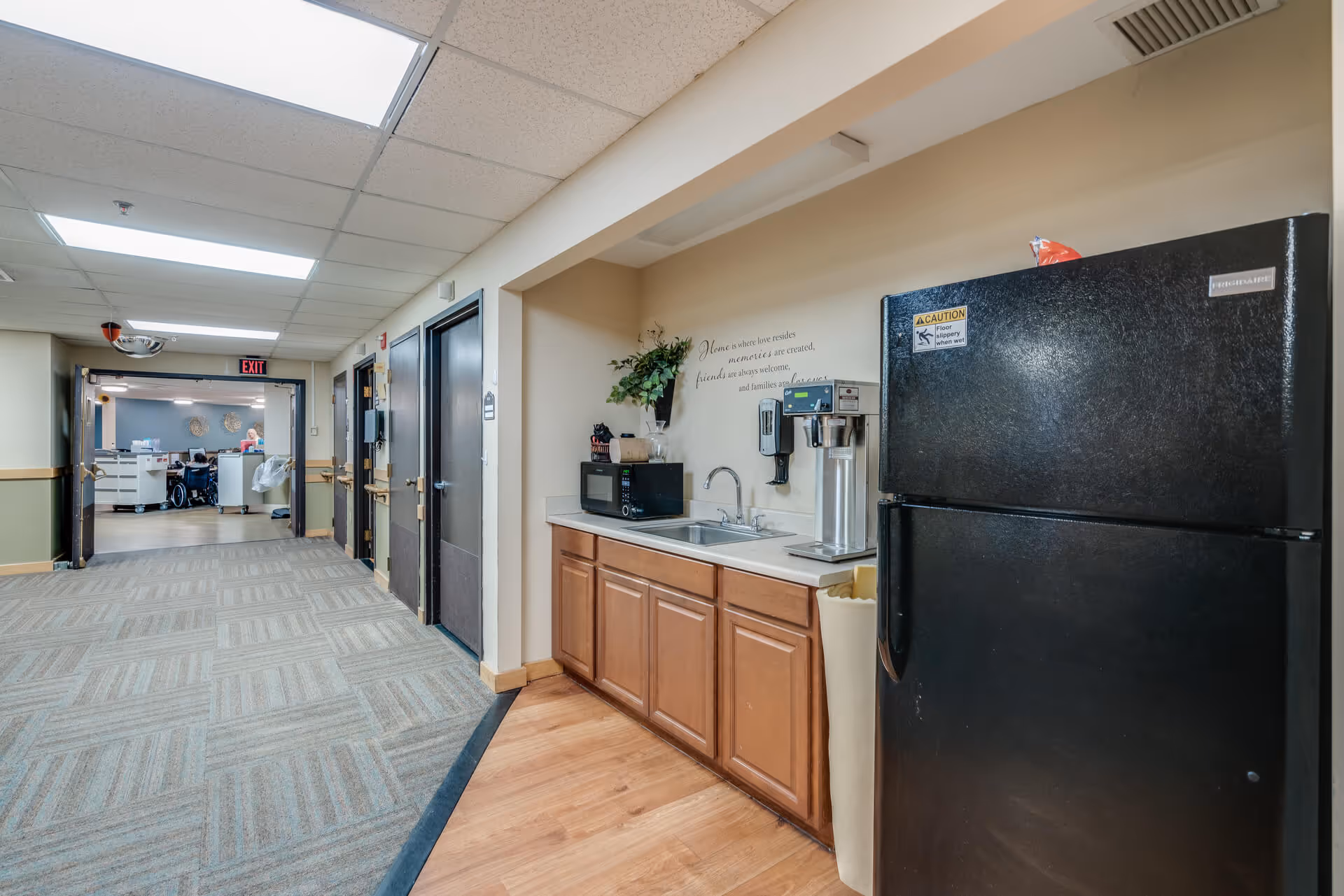 Hallway inside a senior living facility showing a small kitchenette with a black refrigerator, sink and microwave alongside a carpeted corridor leading to a nurses' station.