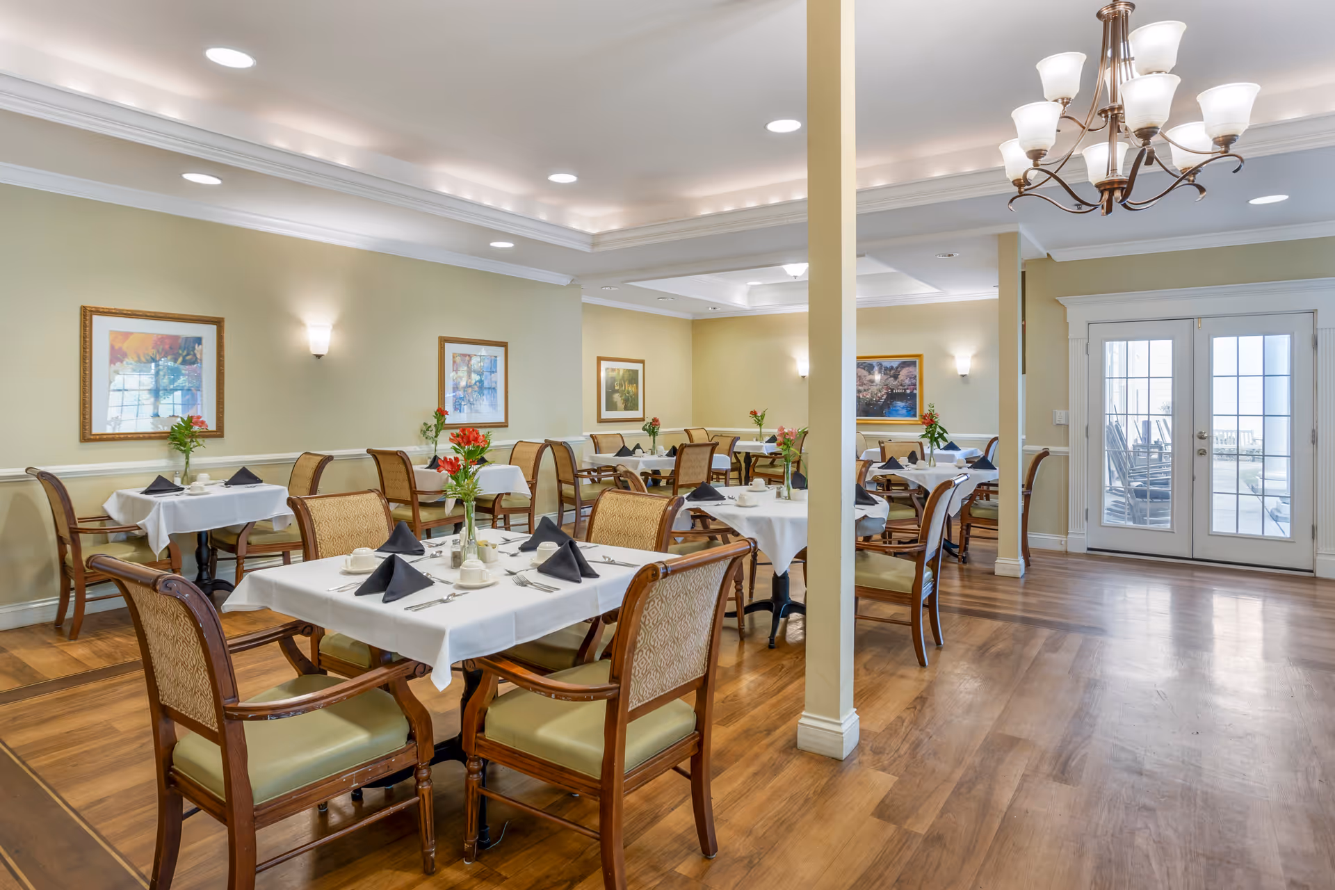 A bright and elegant dining room with several tables covered in white tablecloths, each set with black folded napkins, cups, and silverware. The room features wooden chairs with cushioned seats, framed artwork on the walls, a chandelier, recessed ceiling lights, and double glass doors leading outside.