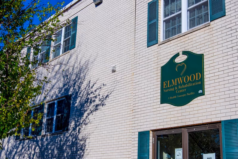 Exterior view of Elmwood Nursing & Rehabilitation Center showing a white brick building with green window shutters and a green sign with gold lettering displaying the facility's name.