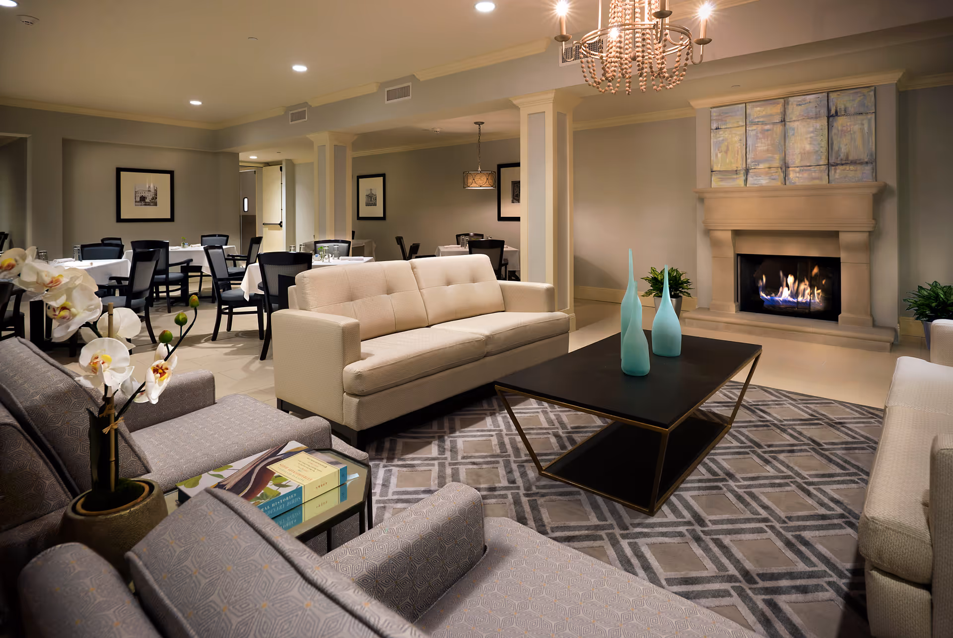 A cozy living room area with beige sofas and patterned armchairs arranged around a black coffee table with two blue decorative vases. A lit fireplace with a large abstract painting above it is on the right wall. In the background, there is a dining area with tables covered in white tablecloths and black chairs. The room is softly lit with recessed ceiling lights and a chandelier.