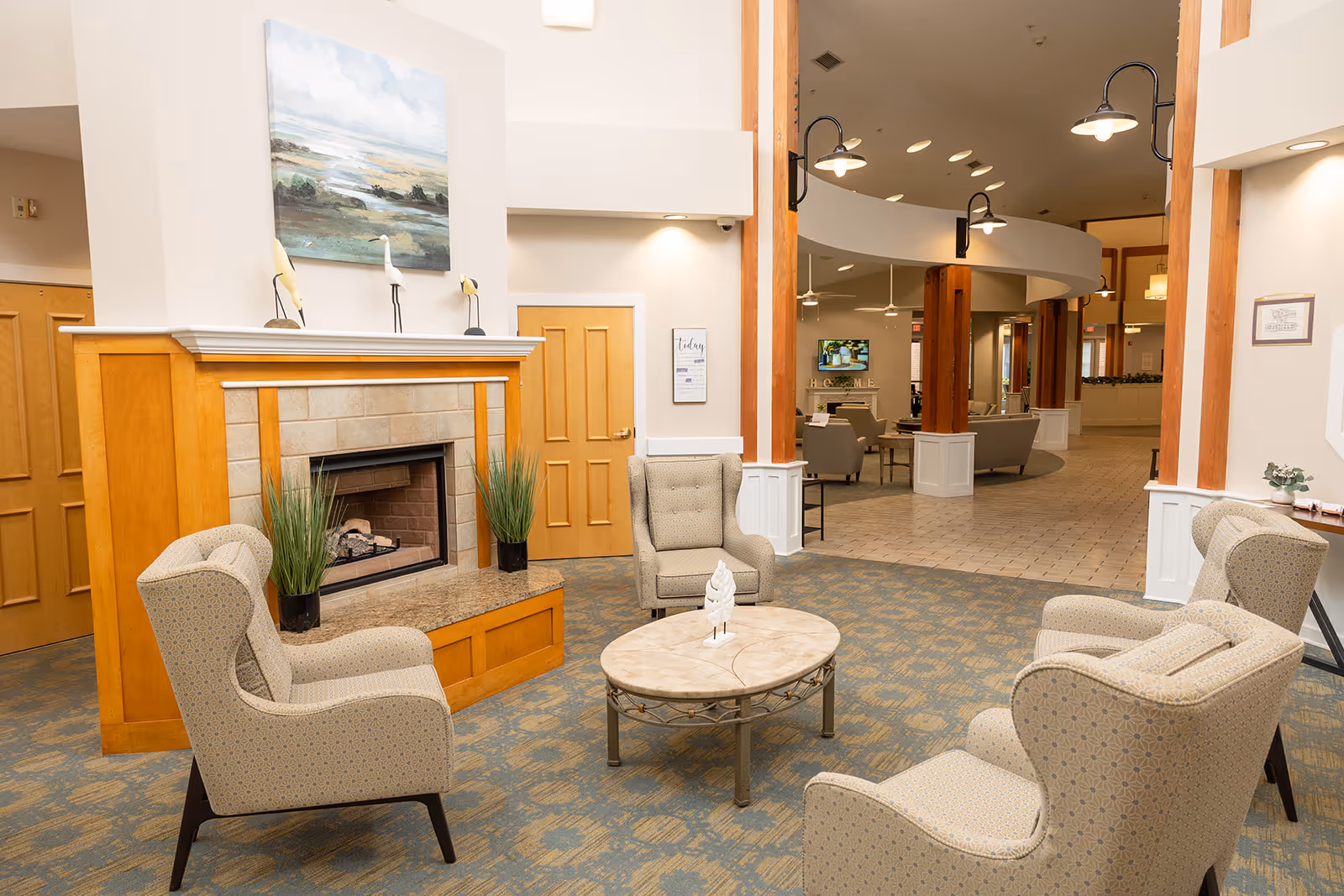 A bright senior living lounge with upholstered armchairs arranged around a round coffee table in front of a wood-paneled fireplace and an open corridor to common areas.