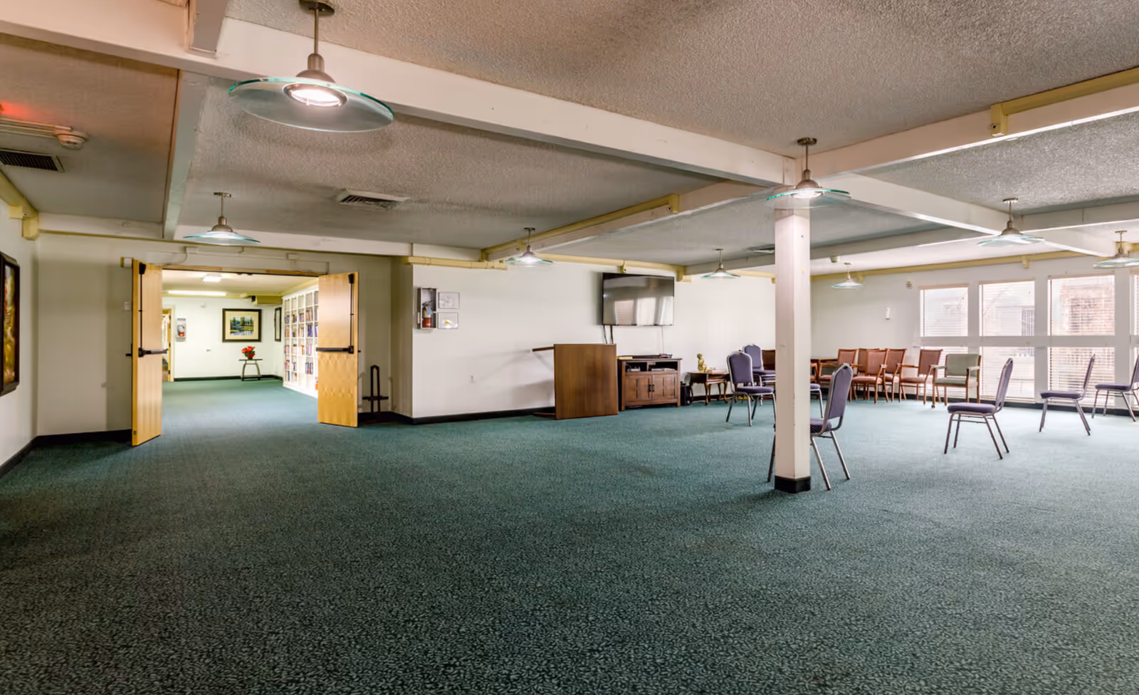 A large, open common room with green carpet and white walls. Several chairs are spaced out around the room, with a few wooden chairs lined up near the windows. The room has a TV mounted on the wall, a wooden podium, and a small table with a decorative item. Ceiling lights hang from the white beams, and double doors lead to another room with bookshelves and framed pictures.