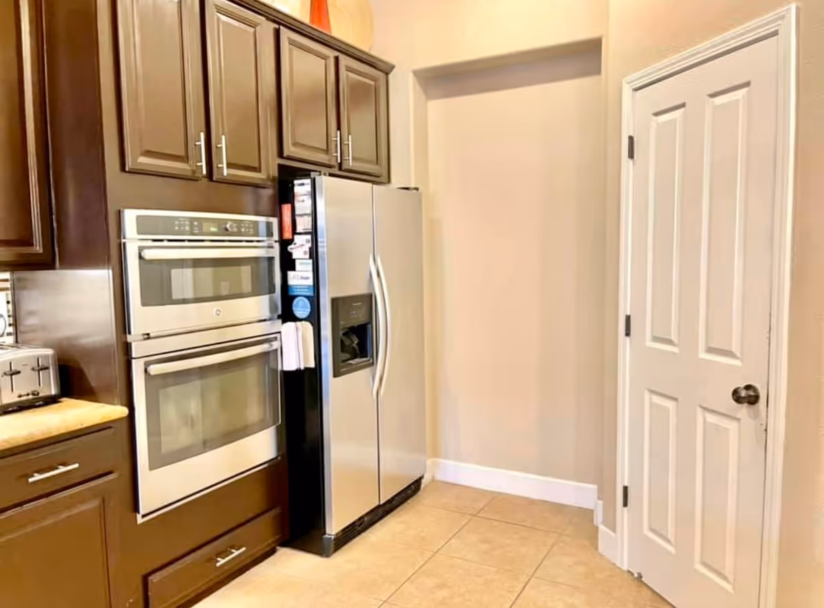 Modern kitchen corner with stainless steel double oven and side-by-side refrigerator. Dark wood cabinets with silver handles are above and below the appliances. Beige tiled floor and light-colored walls with a white door on the right side.