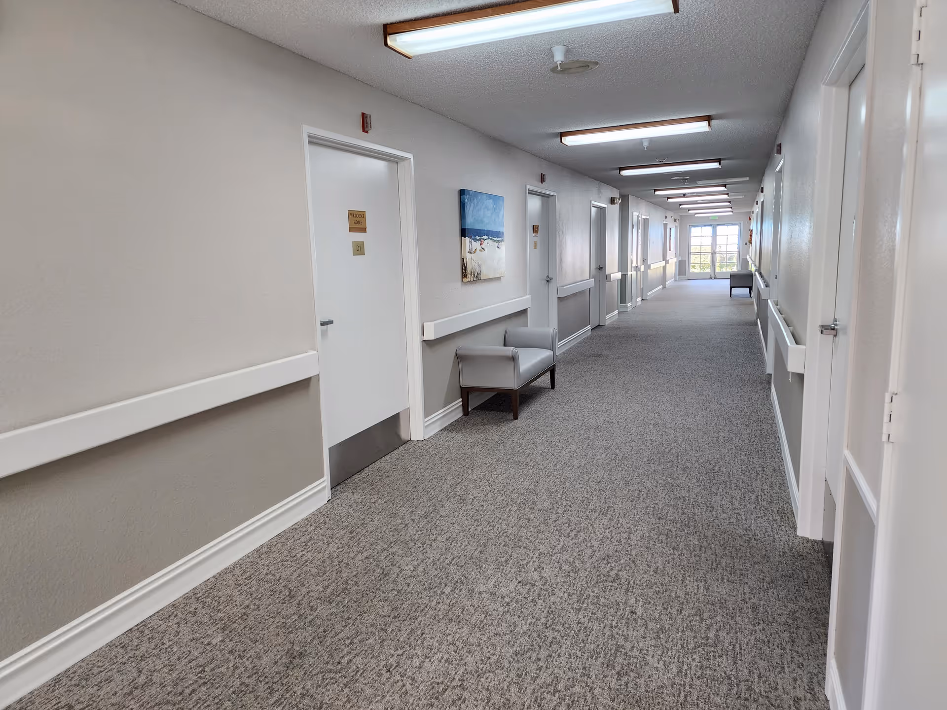 A long, carpeted hallway in a senior living facility with white walls and multiple closed doors on both sides. There are handrails along the walls and a couple of armchairs placed against the walls. A painting depicting a beach scene is hung on the left wall. The hallway is well-lit with ceiling lights and has a window at the far end letting in natural light.