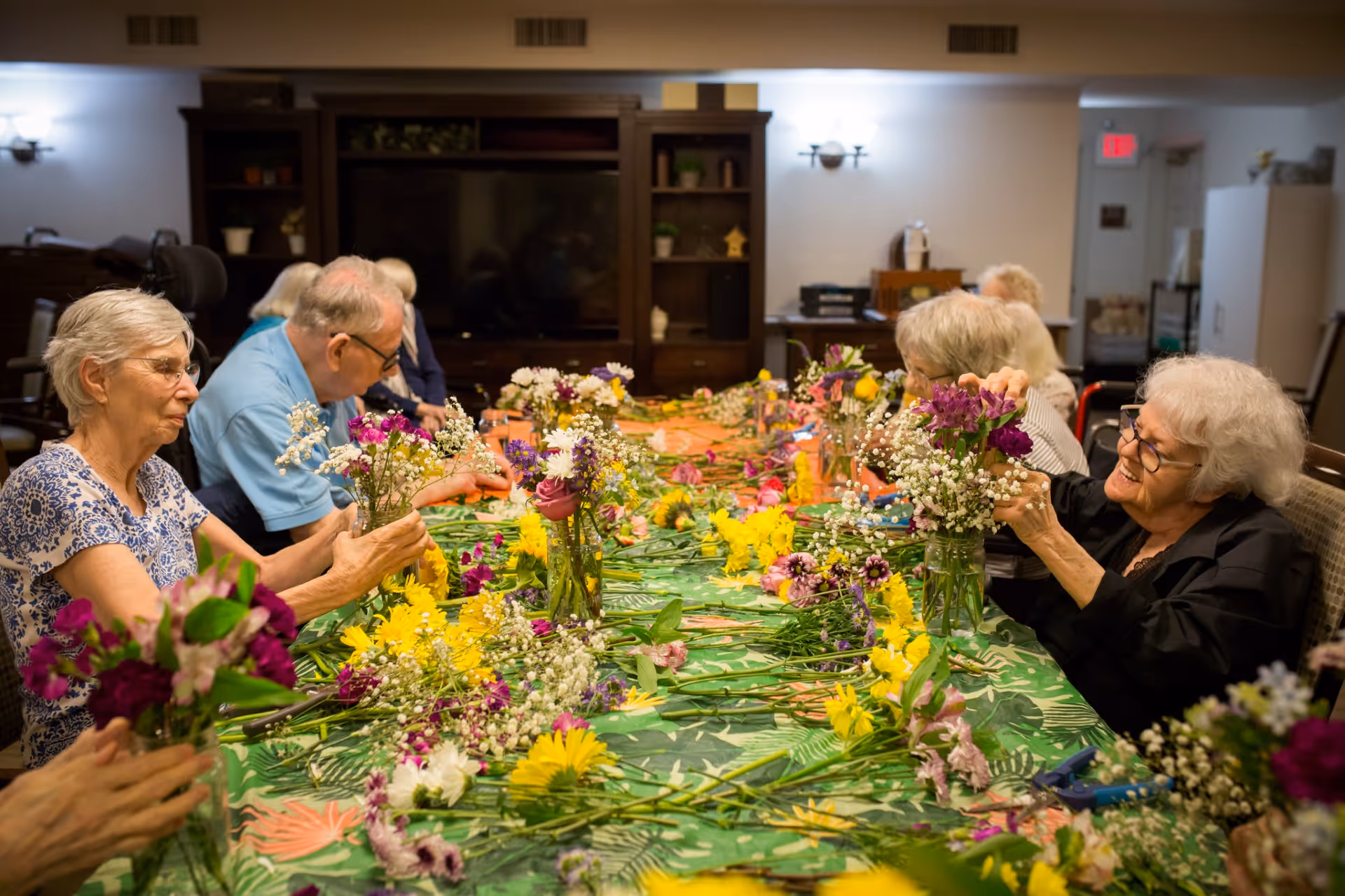 A group of elderly people sitting around a long table covered with a green floral tablecloth, arranging colorful flowers into glass vases in a well-lit room with shelves and a television in the background.