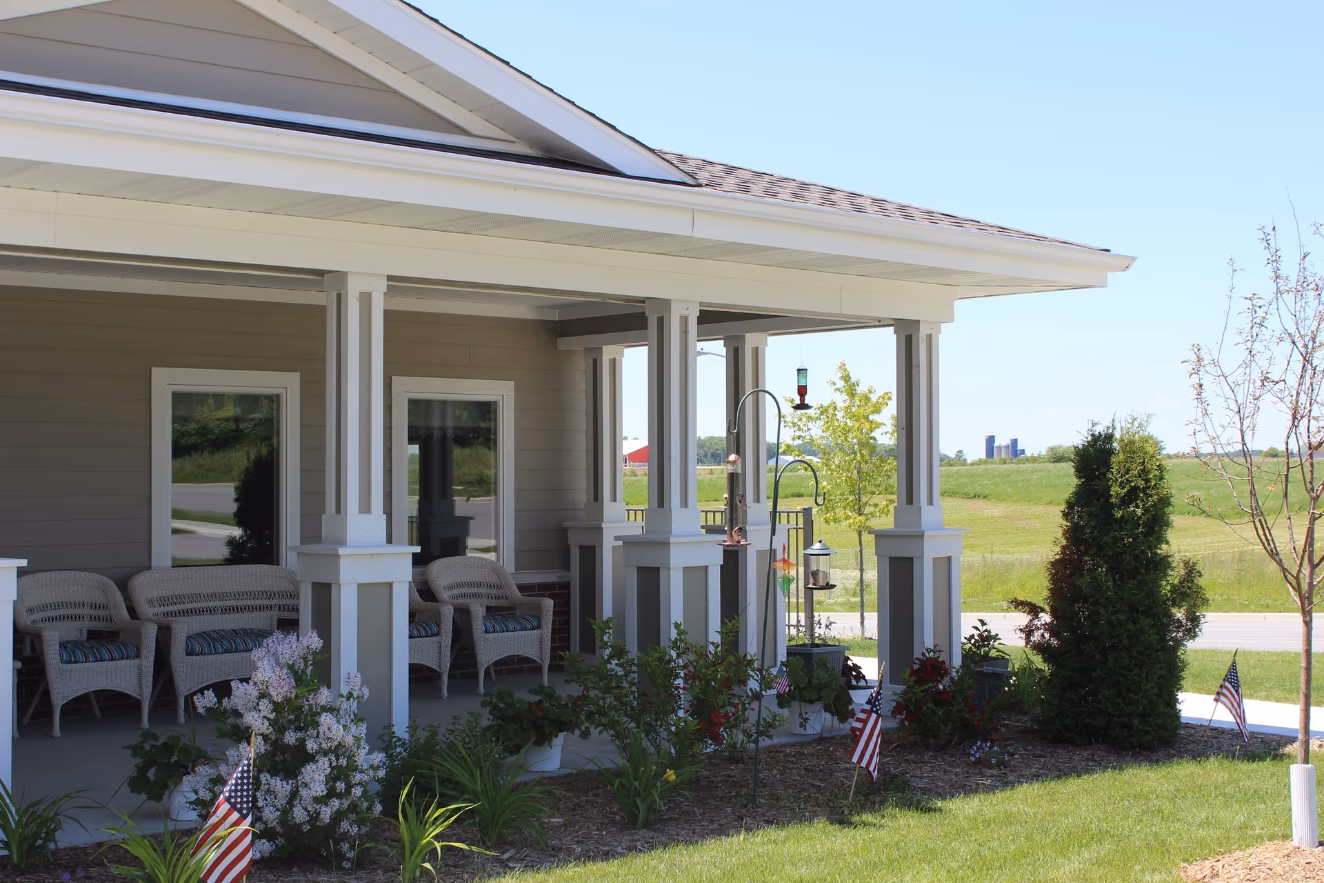 Covered porch area of a senior living facility with wicker chairs and cushions, surrounded by a garden with small American flags and various plants. The porch has white pillars and overlooks a grassy field with a clear blue sky.