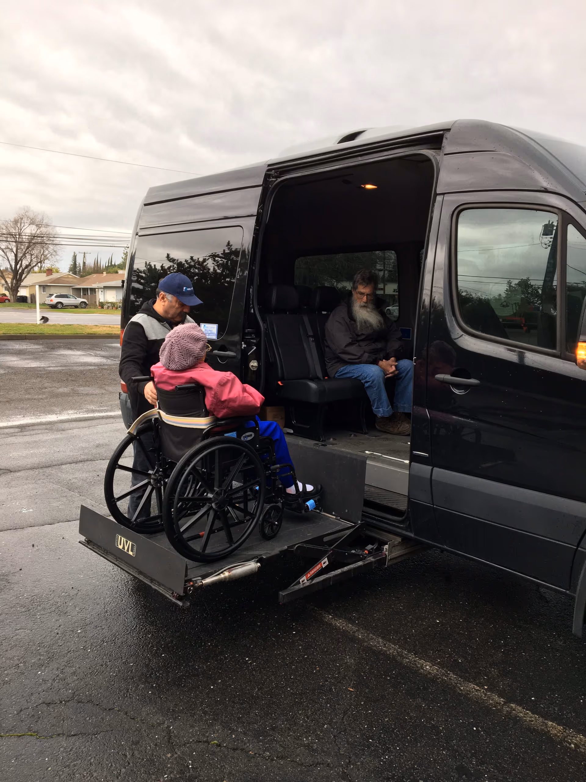 A person in a wheelchair is being assisted by a man onto a black van using a wheelchair lift. Inside the van, an elderly man with a long white beard is seated. The scene takes place outdoors on a cloudy day in a parking lot.