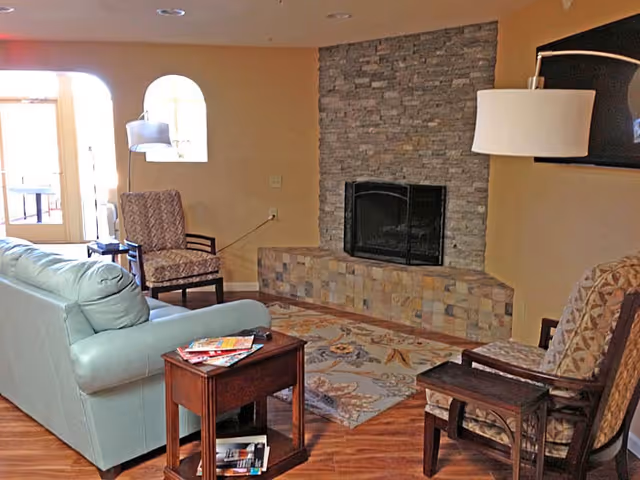 Cozy assisted living lounge with a pale green sofa, patterned armchairs, a stone fireplace, and wood flooring.