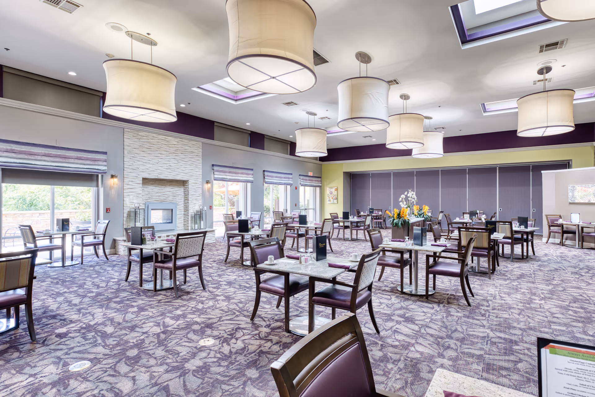 A spacious dining room with multiple tables and chairs arranged neatly. The room features large hanging light fixtures, a patterned carpet, and large windows with blinds allowing natural light to enter. There is a fireplace on one wall and decorative plants on some tables.