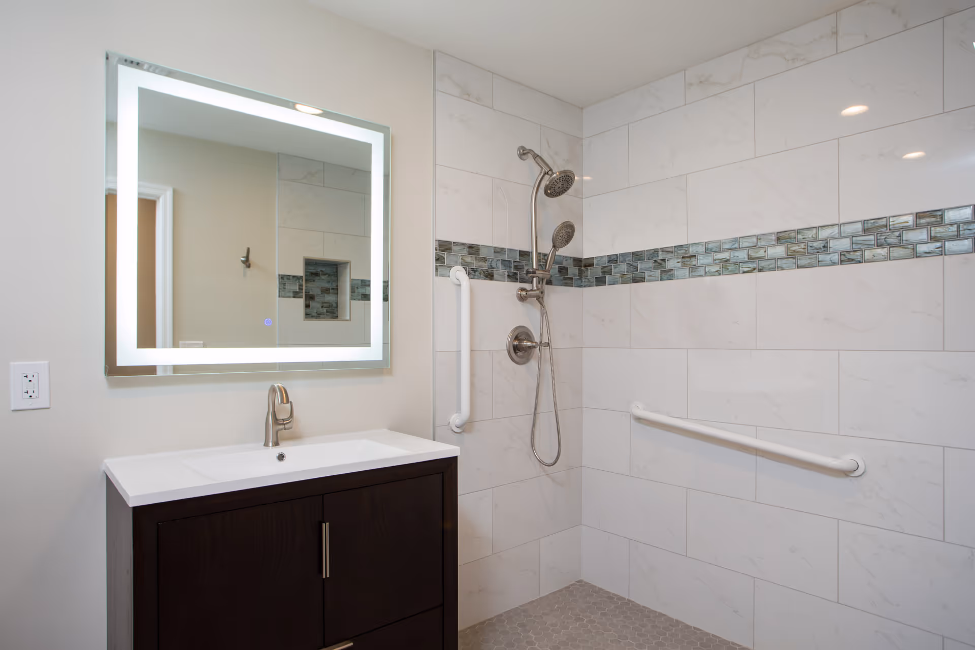A modern bathroom featuring a dark wood vanity with a white countertop and a sleek faucet. Above the vanity is a large rectangular mirror with built-in LED lighting. The shower area has white marble-like tiles with a decorative horizontal strip of blue and green mosaic tiles. There are two shower heads, one fixed and one handheld, along with white grab bars installed on the walls for safety.