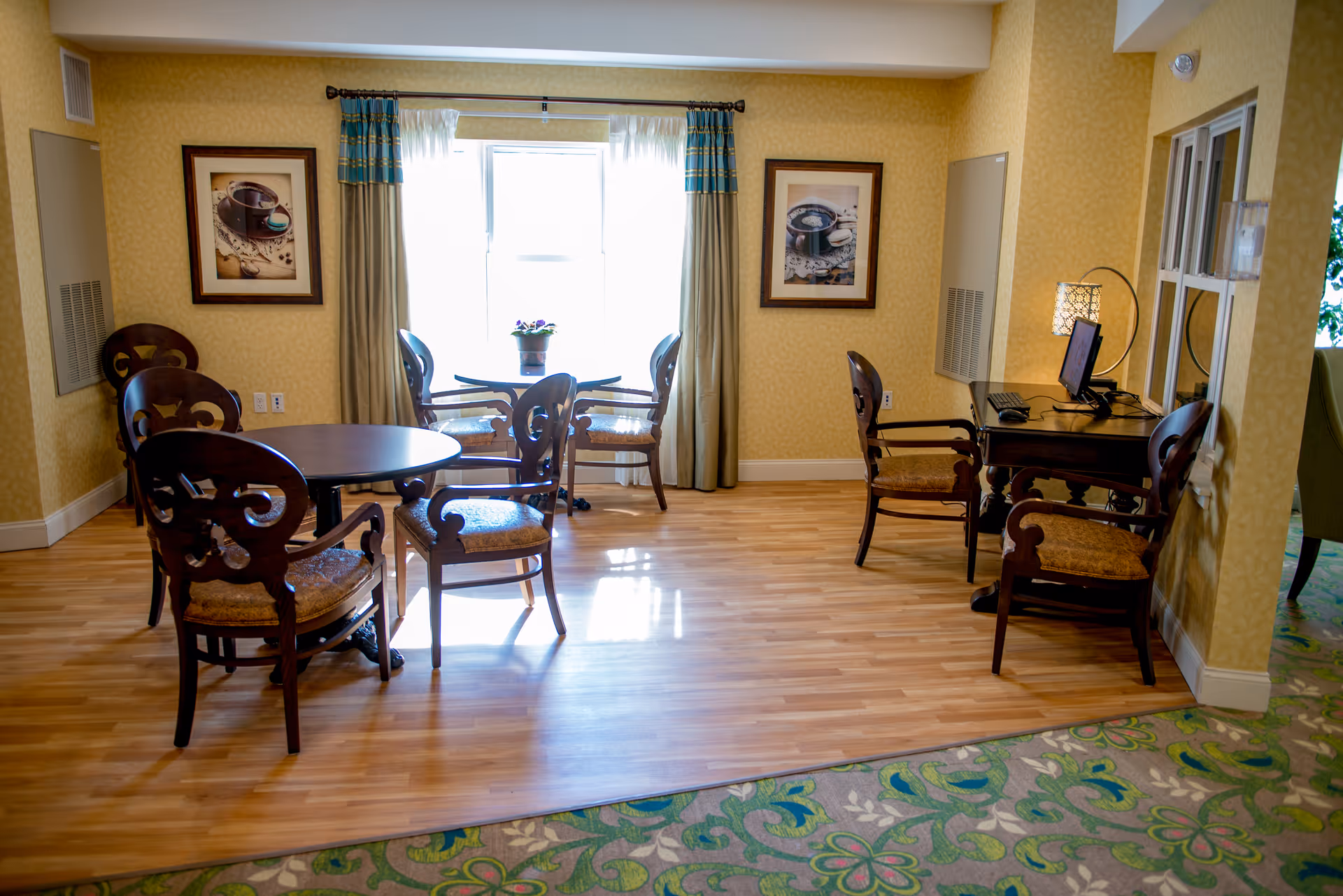 A cozy interior room with wooden flooring and yellow patterned walls. There are two round tables with ornate wooden chairs around them. A window with curtains lets in natural light, and a small potted plant sits on one of the tables. On the right side, there is a small desk with a computer and two chairs. The room has framed pictures on the walls and a green floral carpet partially visible in the foreground.