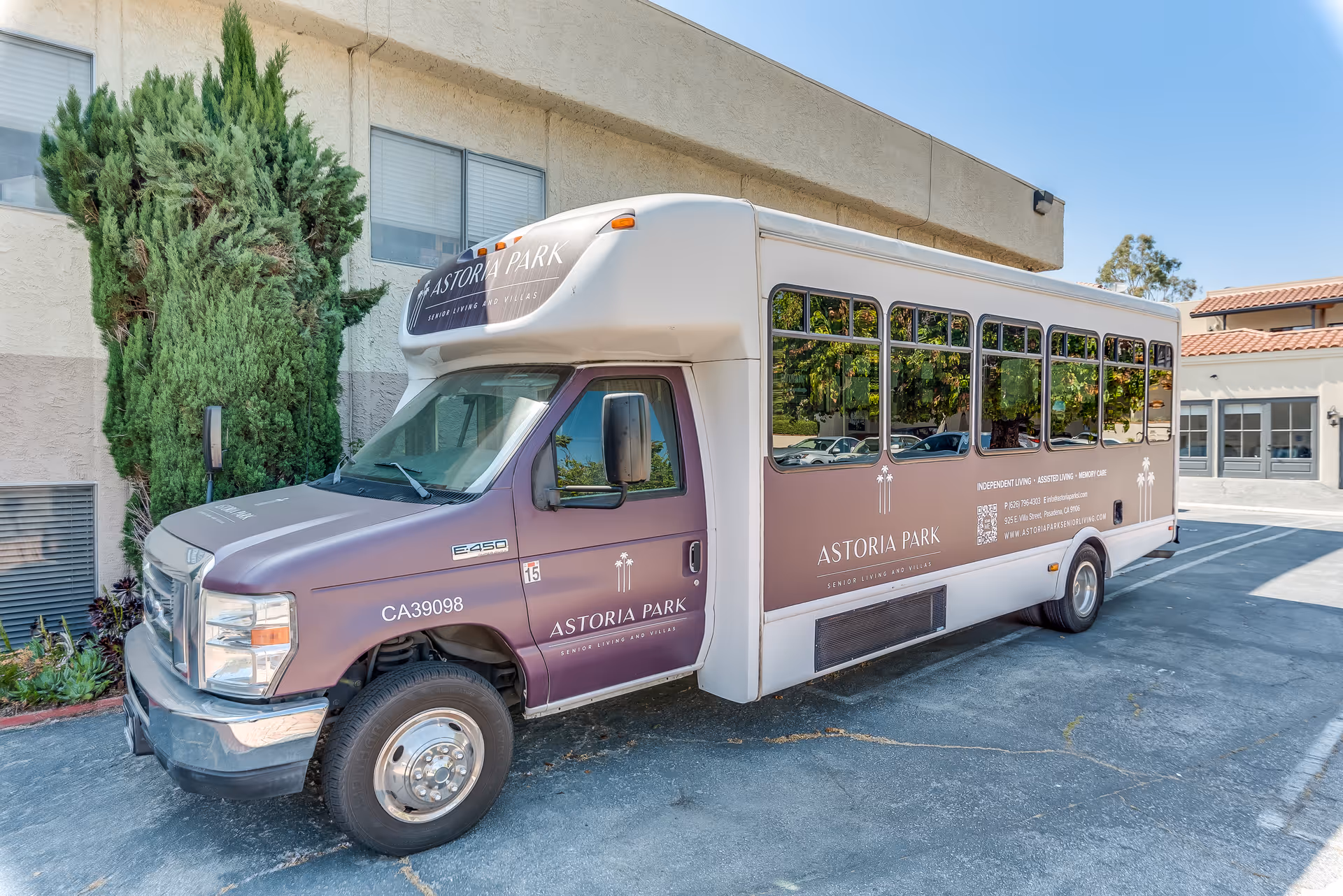 A shuttle bus parked outside a building with the branding 'Astoria Park Senior Living and Villas' on its side. The bus is white and purple with large windows and is parked on a paved area next to some greenery and a beige building.