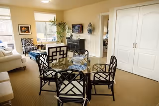 A bright and spacious living area with a glass-top dining table surrounded by four black chairs with geometric designs. The room features beige carpeting, a white sofa, a blue ottoman, a black sideboard with a mounted TV above it, decorative vases, a tall green plant, and large windows letting in natural light.