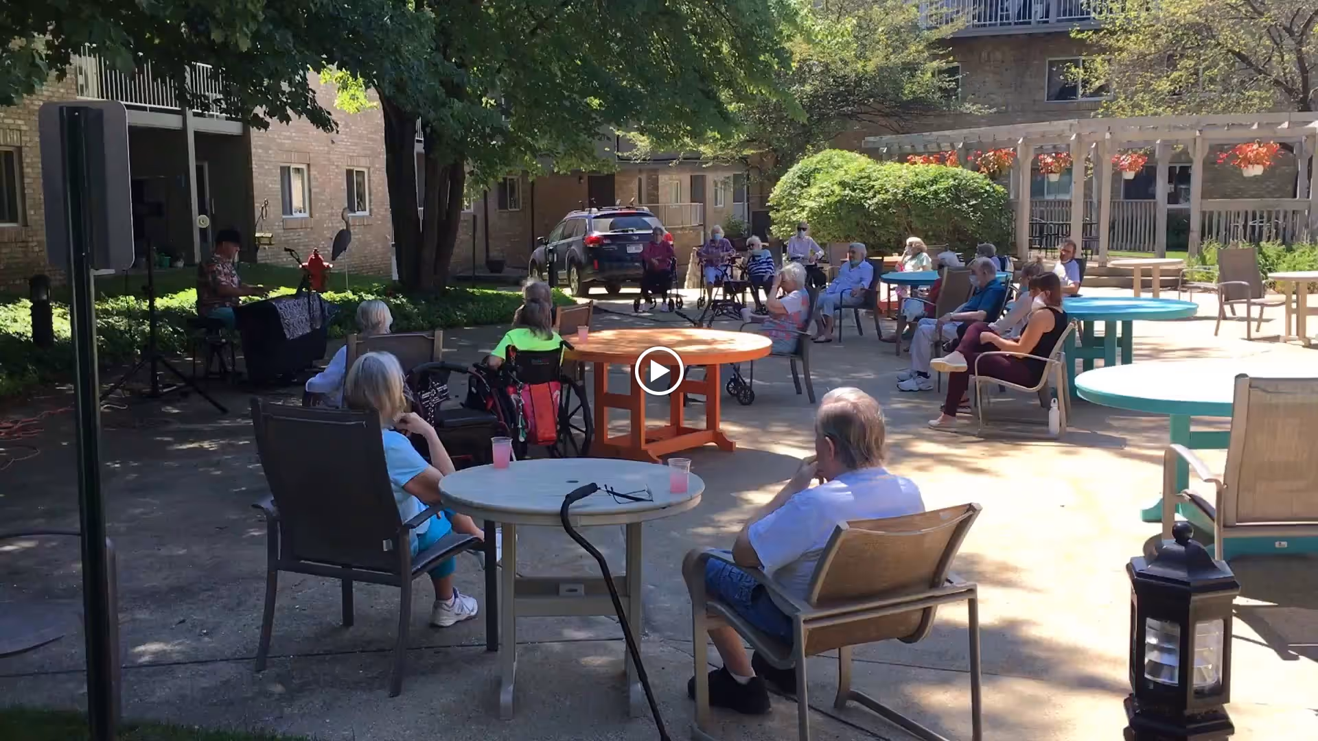 A group of elderly people sitting outdoors at round tables in a courtyard area of a retirement community, listening to a person playing a keyboard under the shade of trees. The setting is sunny with some shaded areas, surrounded by buildings and greenery.