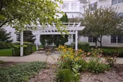 Outdoor garden area with a white pergola, patio tables and chairs, surrounded by green shrubs, flowering plants, and trees, adjacent to a multi-story building with white siding and large windows.
