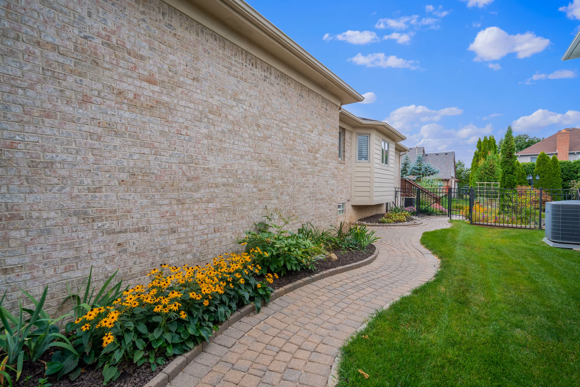 A curved brick pathway runs alongside a beige brick building with a garden bed of yellow flowers and green plants on one side and a well-maintained green lawn on the other. In the background, there is a black metal fence, some trees, and neighboring houses under a partly cloudy blue sky.