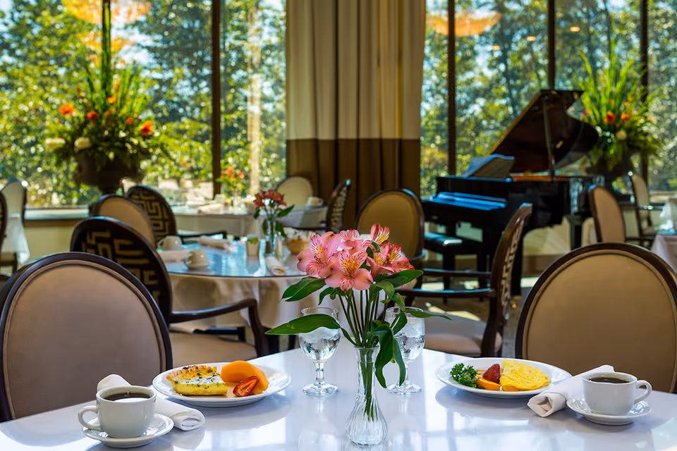 Sunlit dining room with set tables, a vase of pink flowers, breakfast plates and a grand piano by large windows.