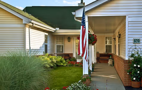 Front porch area of a single-story building with white siding and a green roof. There is a well-maintained lawn with various plants and flowers, an American flag hanging from a white column, and benches along the porch. The building number 32 is visible on the wall.