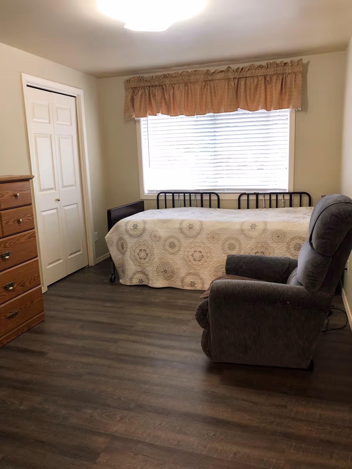 A small bedroom with a single bed covered with a patterned quilt, a gray recliner chair, a wooden dresser with drawers, a window with blinds and a beige valance, and a white closet door.