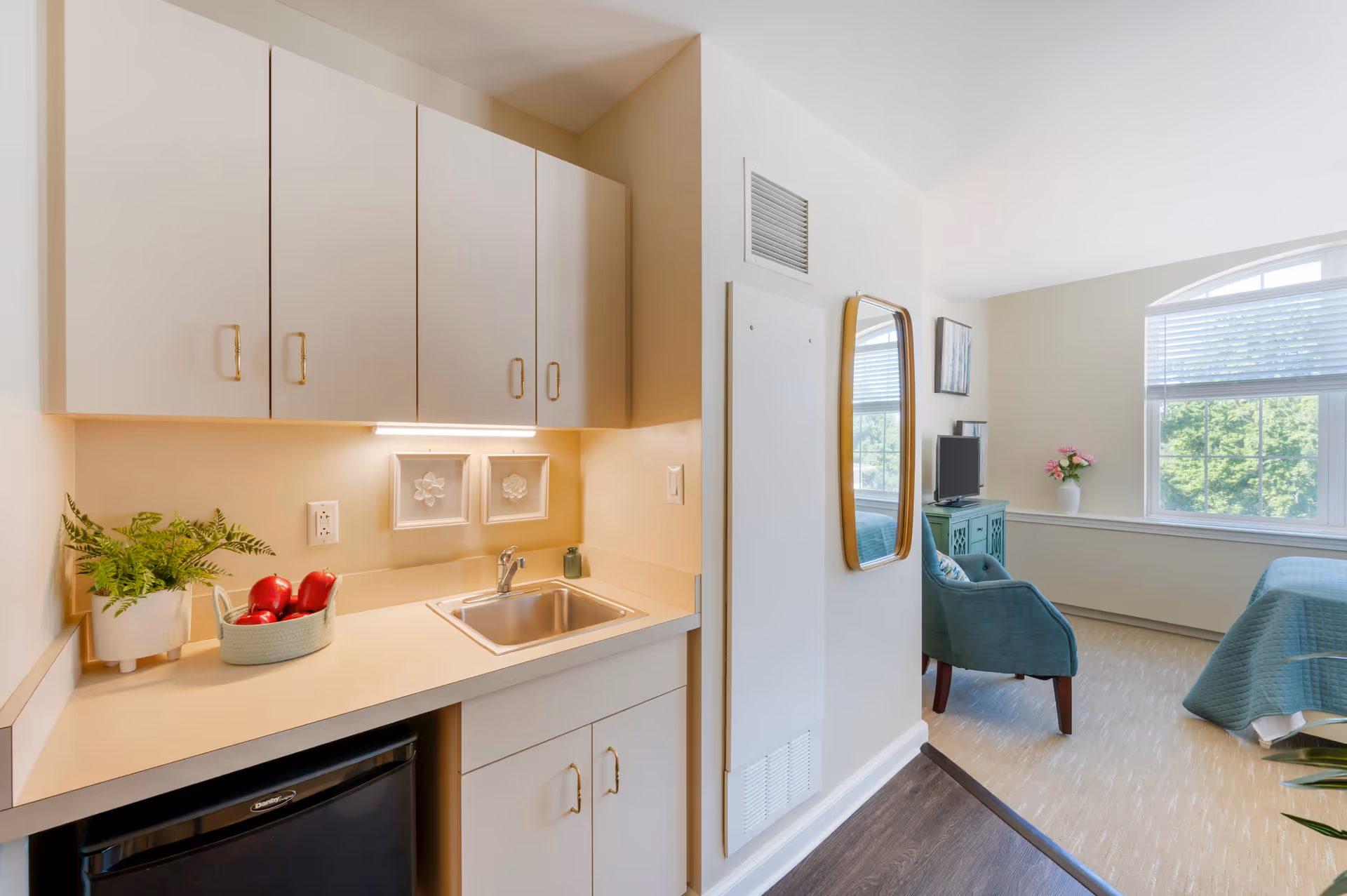 Small kitchenette with sink, cabinets and mini-fridge opening into a furnished bedroom/sitting area with an armchair and bed by a large window.