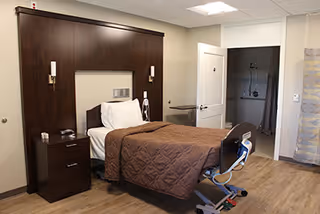 A therapy-style bedroom with a hospital bed, dark wood headboard and nightstand, and an open doorway revealing a walk-in shower bathroom.