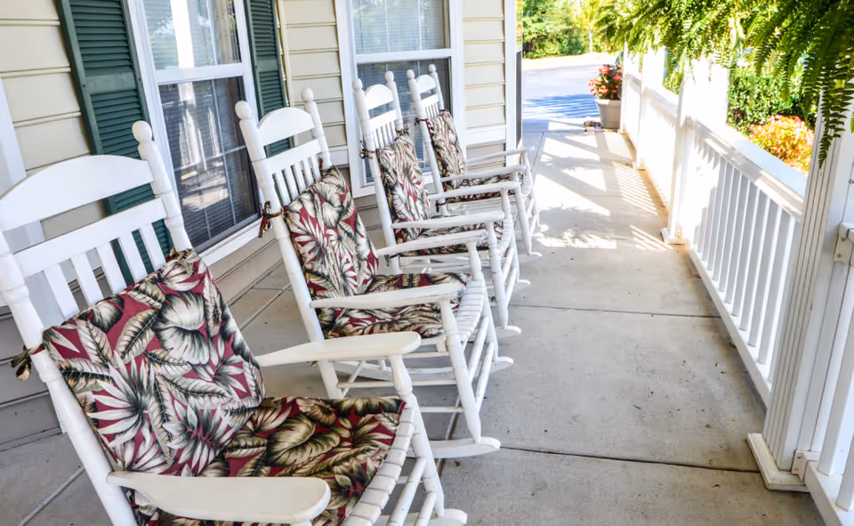 A row of white wooden rocking chairs with floral patterned cushions lined up on a covered porch outside a building with beige siding and green shutters. The porch has a white railing and overlooks a garden area with greenery and flowers.
