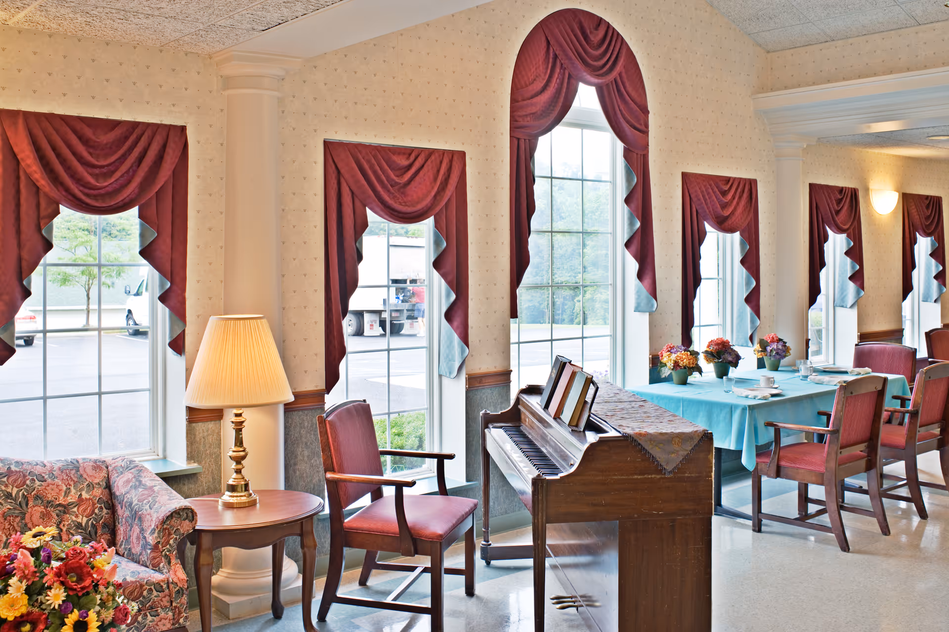 Interior view of a senior living facility room with large windows adorned with red curtains. The room features a floral patterned sofa, a wooden side table with a lamp, a wooden piano with sheet music, and a dining table covered with a blue tablecloth and decorated with flower arrangements. Several wooden chairs with red cushions are arranged around the table.