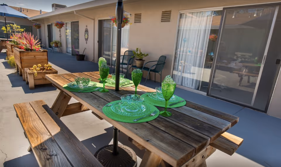 A wooden picnic table set with green plates and glasses on a sunny outdoor patio in front of sliding glass doors and planters.
