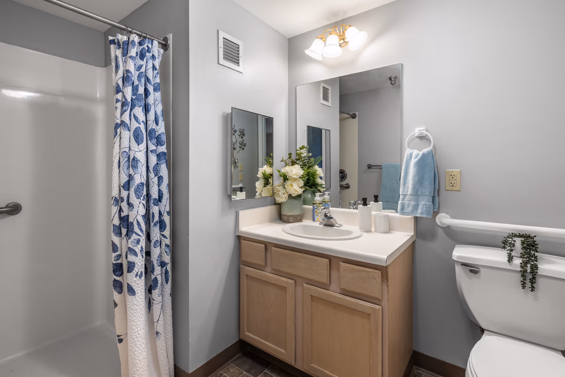A bathroom with a shower area featuring a white and blue leaf-patterned shower curtain, a wooden vanity with a white countertop and sink, a mirror above the sink, a light fixture with three bulbs, a blue hand towel hanging on a ring, and a toilet with a small green plant on the tank.