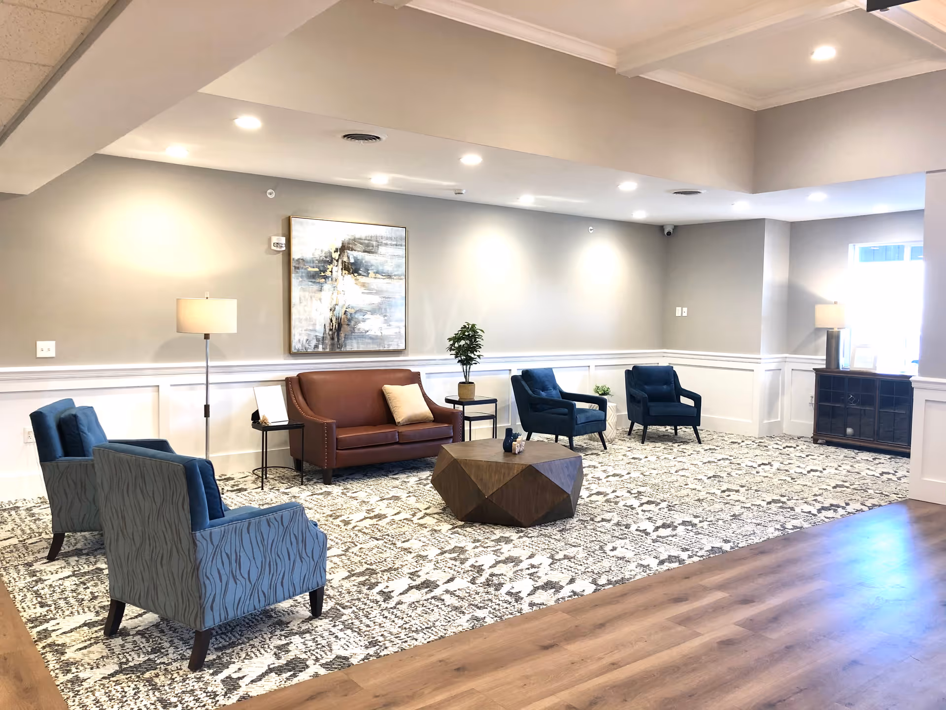 A bright and spacious senior living facility lounge area with a patterned carpet, a brown leather sofa, four blue armchairs, a geometric wooden coffee table, two floor lamps, a side table with a plant, a large abstract painting on the wall, and a wooden cabinet near a window.