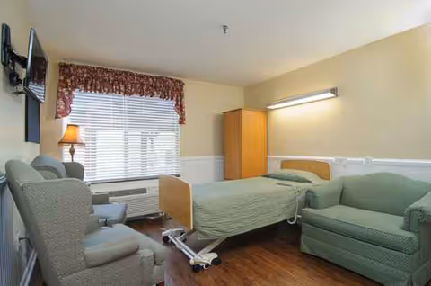 A senior living bedroom with a single bed, green seating area, and window blinds.