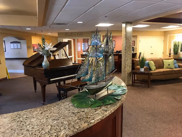 Interior view of a senior living facility lounge area featuring a grand piano with a vase of flowers on top, a decorative model sailboat on a granite countertop, and a seating area with a brown couch and green pillows. The background shows a sign for Shelly's Cafe and warm lighting.