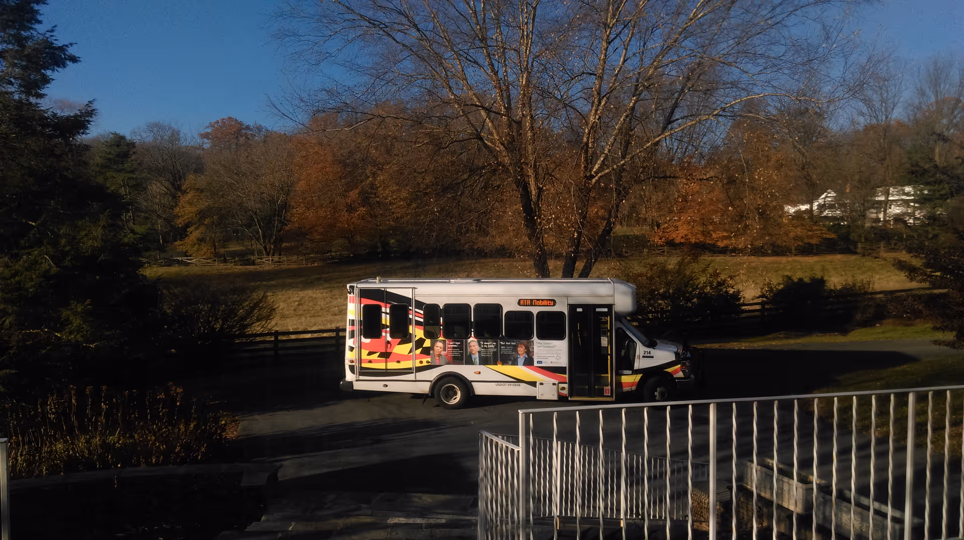 A small shuttle bus parked on a driveway in front of a large tree with autumn foliage. The bus has colorful stripes and images of people on its side. The surrounding area includes a grassy field, trees, and a white fence under a clear blue sky.
