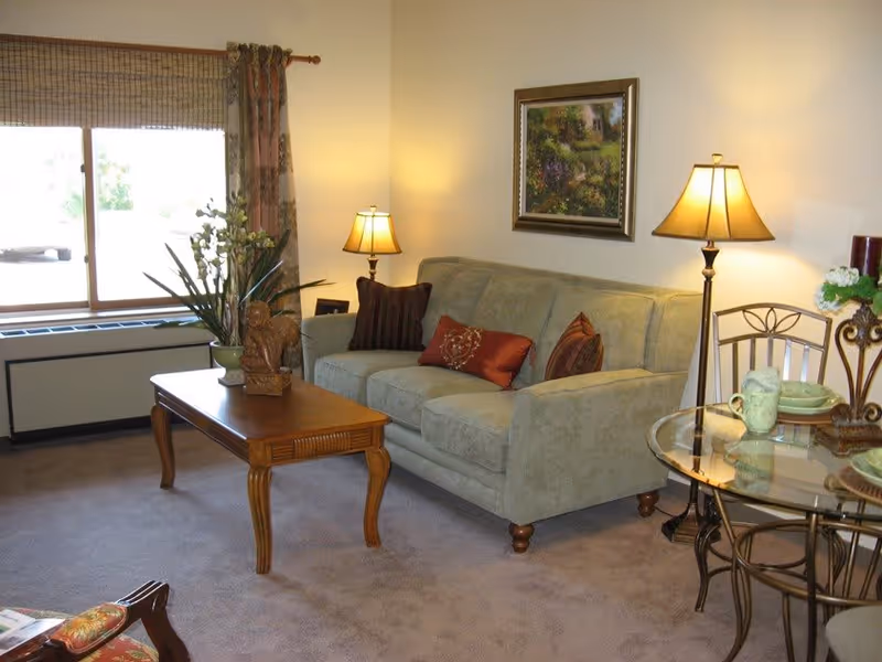 A cozy living room with a light green sofa adorned with decorative pillows, a wooden coffee table with a plant and sculpture, two floor lamps with warm lighting, a framed painting on the wall, a window with curtains, and a glass-top dining table with chairs and tableware.