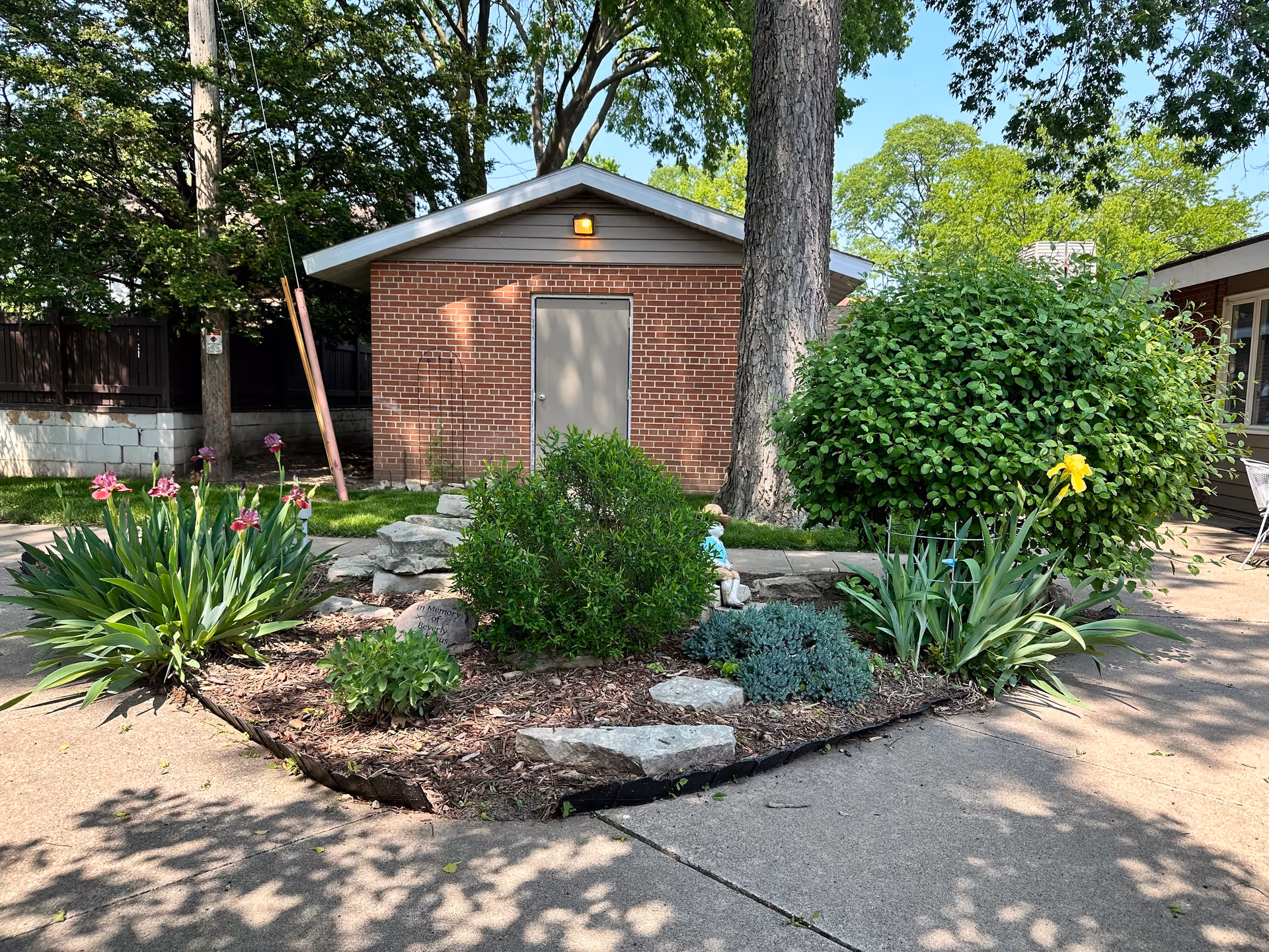 A small garden area with various plants and flowers, including pink and yellow blooms, bordered by a concrete walkway. Behind the garden is a brick building with a closed gray door and a light above it. Trees and bushes surround the area, providing shade and greenery.