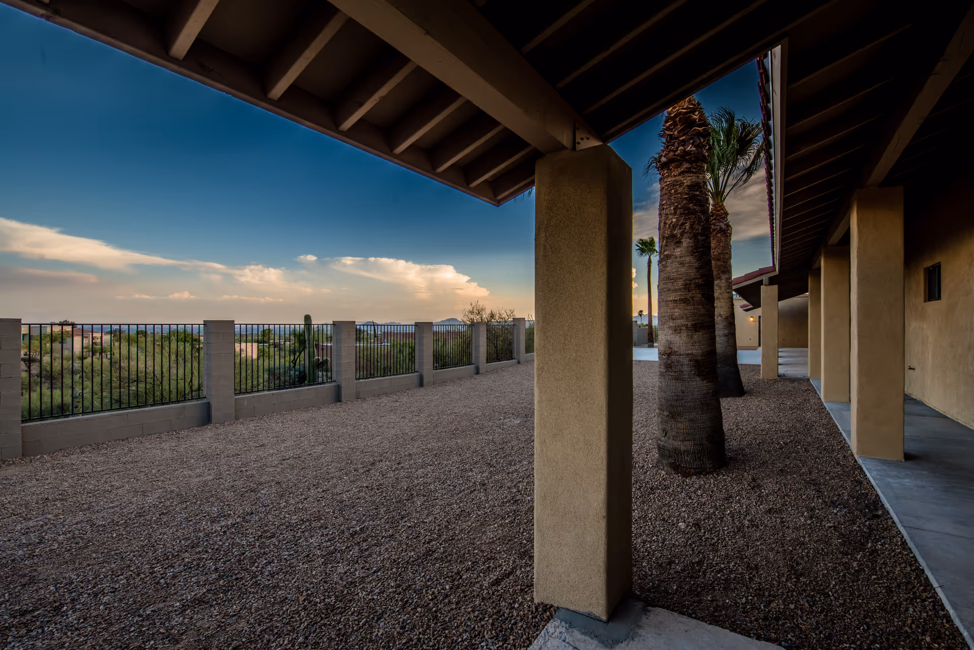 Covered exterior walkway with stucco columns and palm trees overlooking a gravel yard and metal fence at sunset.