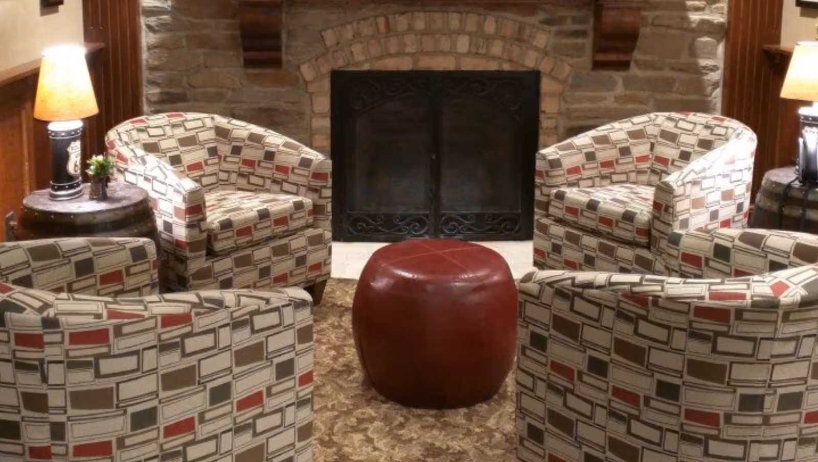 Four patterned armchairs arranged around a red ottoman in front of a stone fireplace with side tables and lamps.