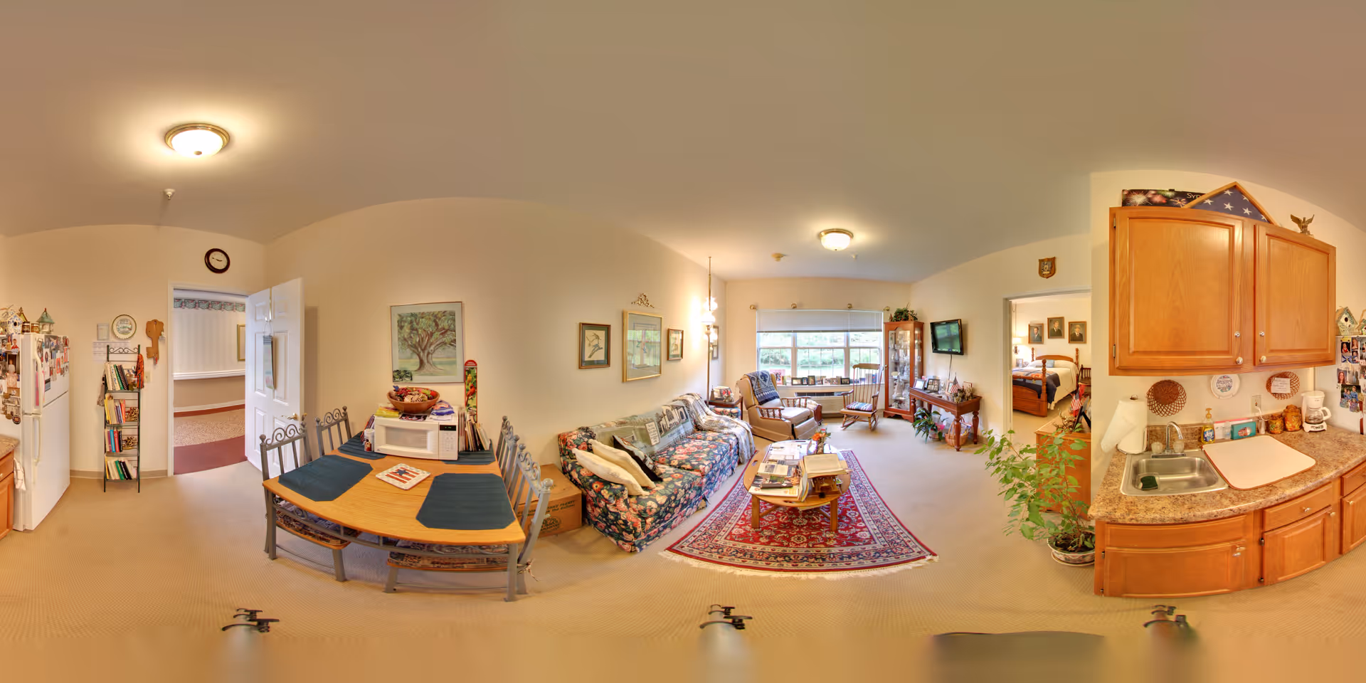A panoramic view of a cozy senior living facility interior at Morningside of Auburn, showing a combined living and dining area with a small kitchen. The dining area has a wooden table with four chairs and placemats. The living room features a floral-patterned sofa, armchairs, a coffee table on a red patterned rug, and a large window letting in natural light. The kitchen area has wooden cabinets, a countertop with a sink, and various kitchen items. The walls are decorated with framed pictures and there are plants adding greenery to the space. A doorway leads to another room with a bed visible.