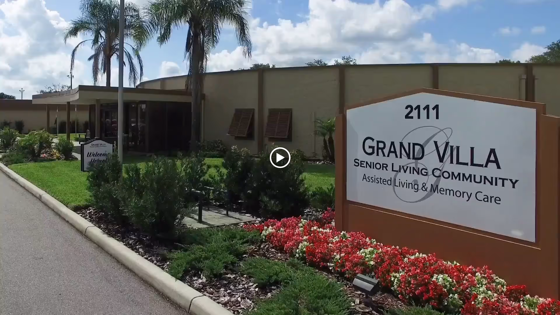 Exterior view of the Grand Villa Senior Living Community building with a sign displaying the address 2111 and information about assisted living and memory care. The scene includes a driveway, landscaped garden with flowers and shrubs, and palm trees under a partly cloudy sky.