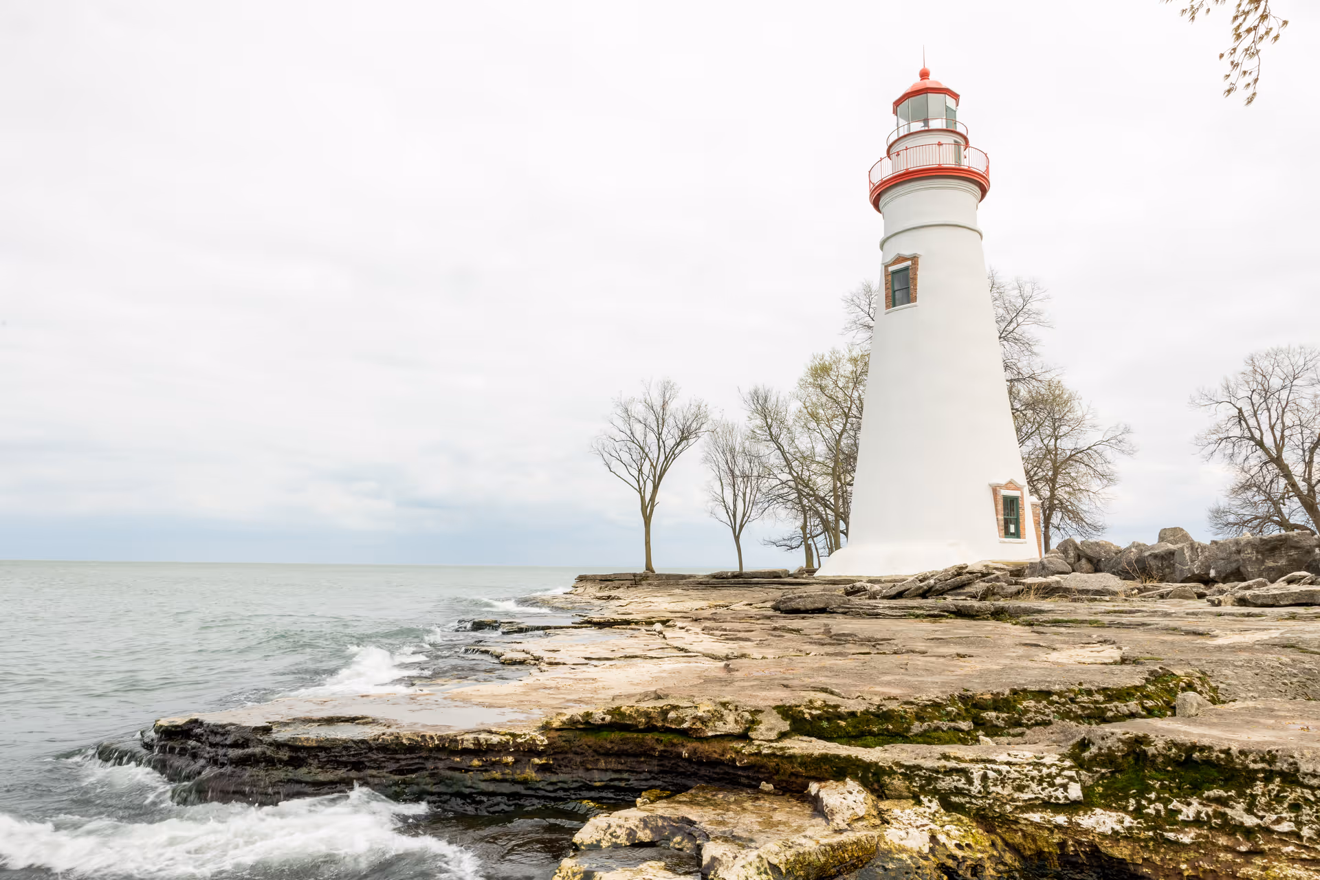 A white lighthouse with a red top stands on a rocky shoreline next to a body of water under a cloudy sky. There are a few leafless trees near the lighthouse.