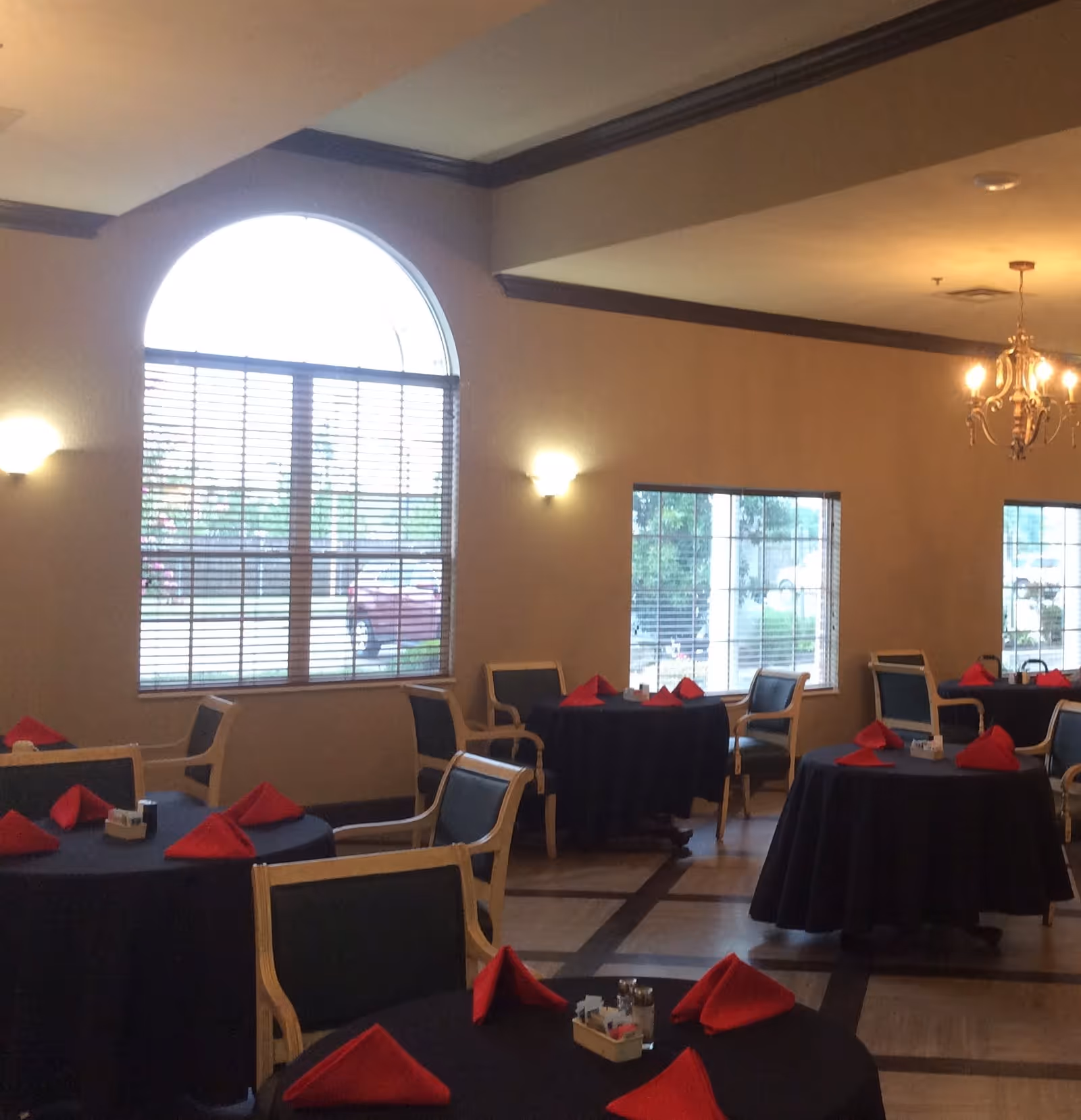 Interior view of a dining room with round tables covered in black tablecloths and red folded napkins. Each table is surrounded by wooden chairs with dark cushions. Large windows with blinds allow natural light to enter the room. A chandelier hangs from the ceiling, and wall sconces provide additional lighting.
